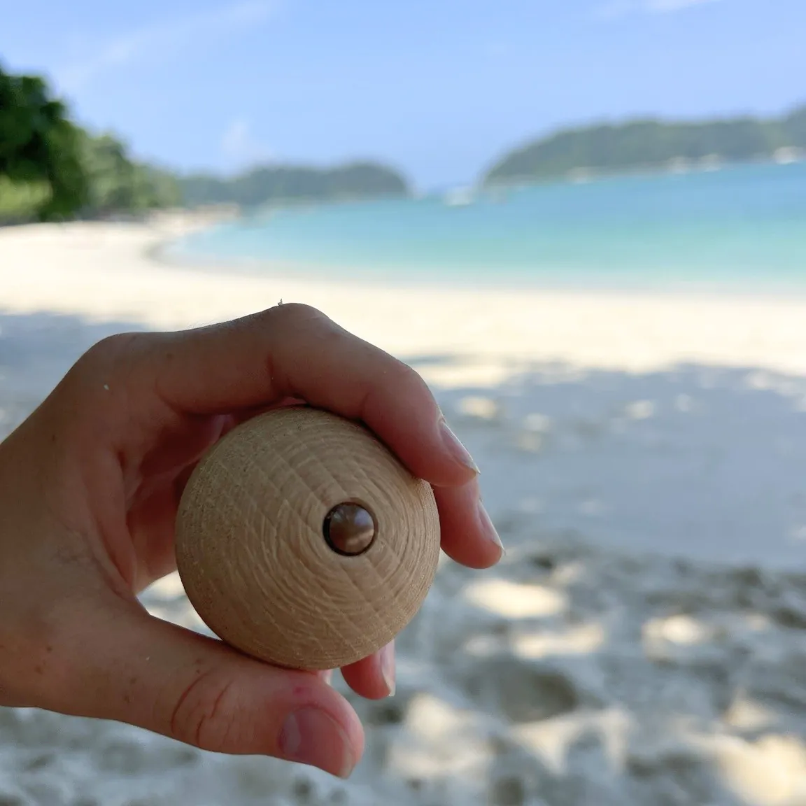 A hand holding the ball toward the camera, a blurred white beach and turquoise sea behind
