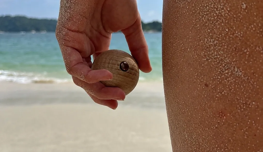 A hand holding a small beech wood ball with a copper core above a sandy beach, the sea behind