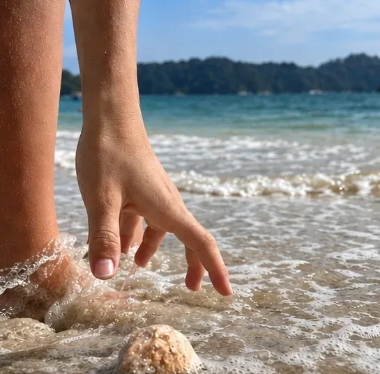 A hand reaching down to touch the sea foam at the water's edge